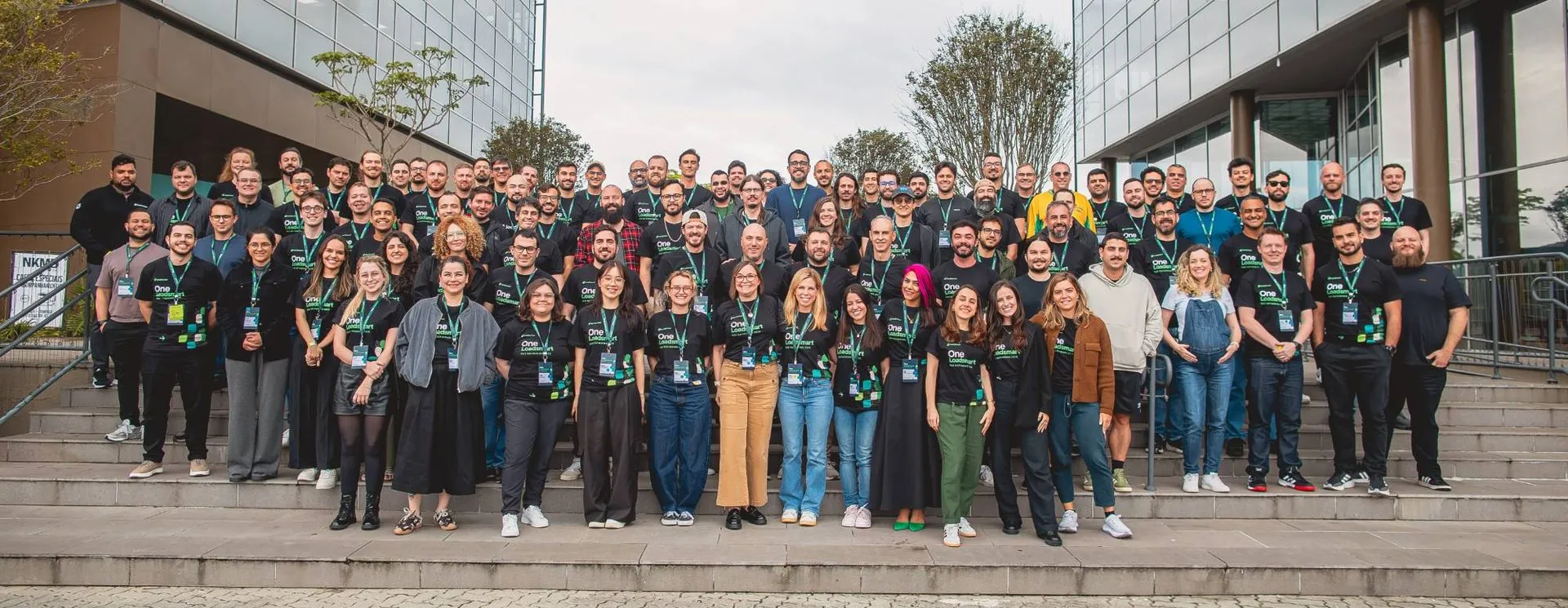 The Loadsmart team, more than a hundred people in black One Loadsmart shirts, gathered on the steps of a glass office building